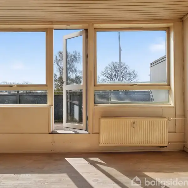 Værelse med åbne dobbeltdøre fører til en balkon. Trægulve, radiator under vindue, og udsigt til træer udenfor. Loftet er af træpaneler.