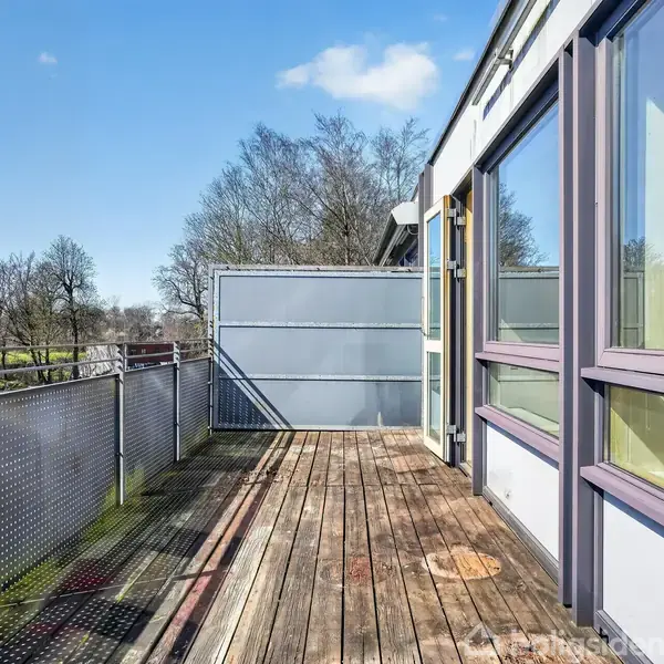 Balkon i træ er tom og solbelyst. Glasskydedøre åbner mod udendørsområdet. Omgivet af træer og blå himmel.
