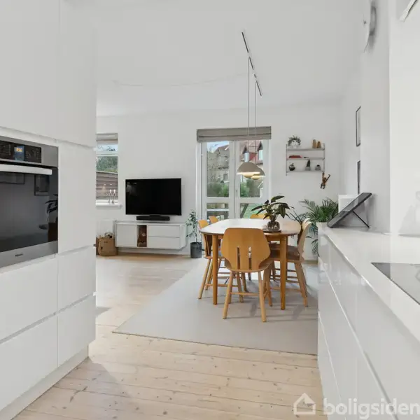 Et moderne køkken-alrum i hvide nuancer indeholder indbyggede apparater til venstre og en kogeplade til højre. Langbord med stole står midt i rummet. Stuen er i baggrunden.