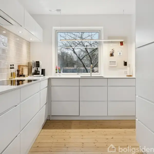 Et moderne køkken med hvide skabe og skuffer; en ovn til højre. Et vindue med udsigt til træer, og en lys armatur hænger over en træbordplade. Hvid flisevæg bagved.