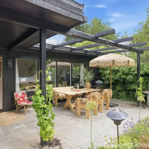 En overdækket terrasse med et træbord og stole, dekorativ parasol og udendørs grill. Omgivet af grønne buske og træer med et kig ind i huset gennem åbne døre.