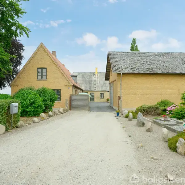 Gårdhus står ved en grusvej omgivet af buske og træer; det har en trekanthusgavl og gulmalet facade. En stensti fører op til indgangen.