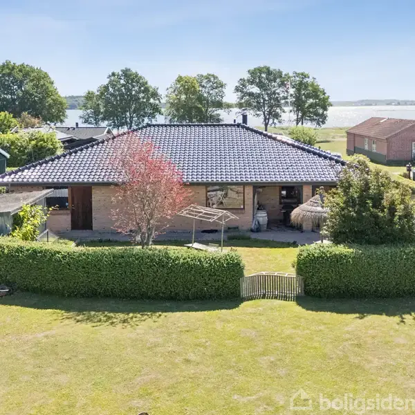 Hus med sort tegltag omkranset af hæk og græsplæne. Det står i et naturskønt område med udsigt til vand og andre huse i baggrunden.