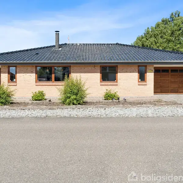 Et enkeltstående hus med gult murværk står stille foran en flisebelagt gårdsplads omgivet af lave buske. Grå tagsten pryder taget, og en garage er i højre side.