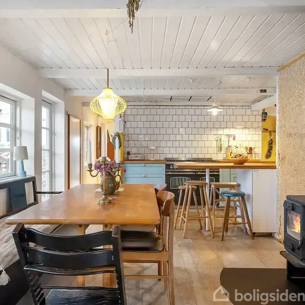 Et rustikt køkken og spisestue med et langbord omgivet af stole. En brændeovn står til højre, mens et oplyst køkkenområde med hvide fliser og barstole ses i baggrunden.