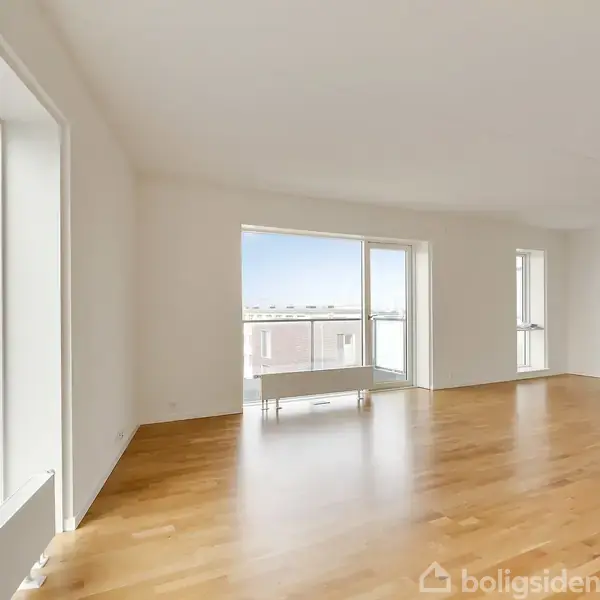 Stort, lyst rum med trægulv strækker sig ud mod store vinduer, der vender mod en balkon med udsigt. Ingen tekst i billedet.