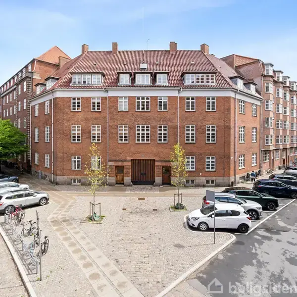 En rød murstensbygning står stille på et gadehjørne omgivet af parkerede biler og cykler. Klare blå himmel danner baggrund, mens træer tilføjer grønne nuancer til bymiljøet.