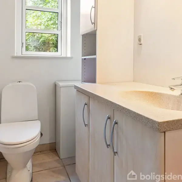 Toilet ved siden af hvidt skab. Håndvask med blandingsbatteri indbygget i skab. Lille vindue i baggrunden giver udsigt til grønt. Flisegulv og hvidmalede vægge.
