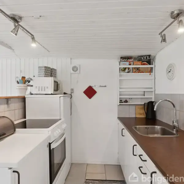 Køkken med hvidmalede vægge og loft, indeholder komfur, mikroovn på hylde, vask, og åbne hylder med bøger. Loftet har spotbelysning og væggen en ur. En rød klud hænger på en krog.