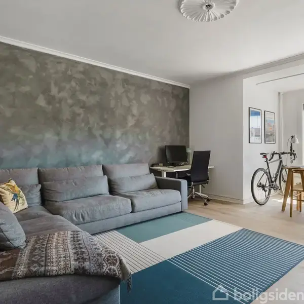 Grå sofa står i en moderne stue med dekoreret væg. Ved siden af ses et skrivebord og en computer, og en cykel står nær spisebordet ved et stort vindue.
