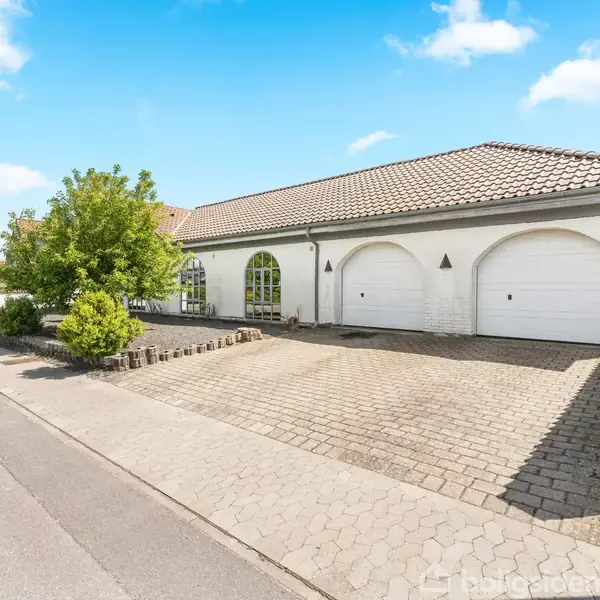 Hvid bygning med to store, buede garageporte står stille ved en flisebelagt indkørsel. En velplejet have med træer sidder ved siden af. Klar himmel og betongade omkranser området.