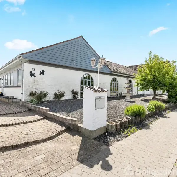 Et hvidt hus med muret facade står på en flisebelagt gade. Nummer 12 er angivet ved siden af døren. En velplejet have omkranser huset, og lampen lyser indgangen.