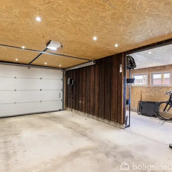 Garage med åben dør til tilstødende rum. Garageporten er lukket, og væggene er delvist træpaneler. I det tilstødende rum ses en cykel og opbevaringshylder. Loftet har indbyggede spotlys.