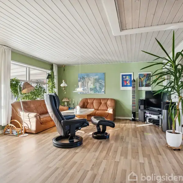 En stue med trælaminatgulv og hvidt træloft. To lænestole og sofa i brun læder omkranser et sofabord. Grønne planter og kunst på væggene skaber en indbydende atmosfære.