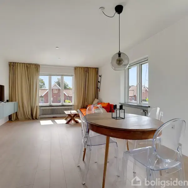 En lys stue med et rundt spisebord og transparente stole. Et tv er vægmonteret til venstre, og en sofa med orange puder står ved vinduet med beige gardiner.