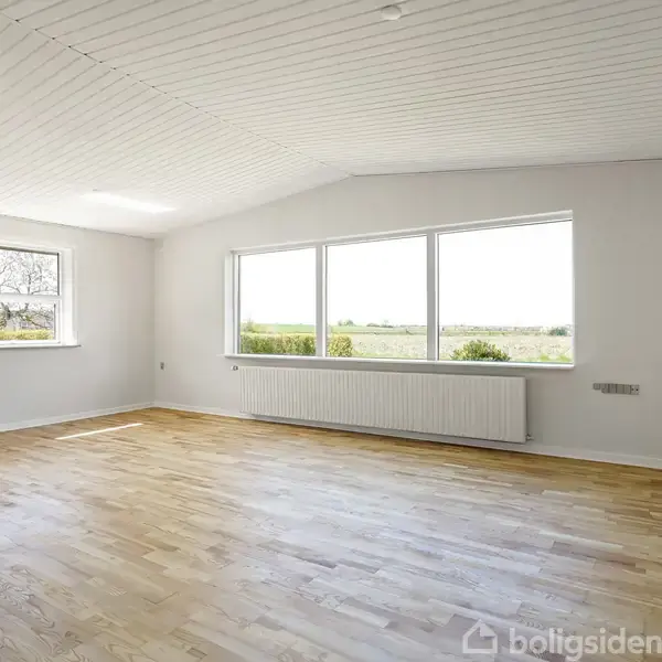 Et tomt rum med parketgulv har to store vinduer, der giver udsigt til en grøn have. Rummet har hvide vægge og et hvidt loft med indbygget radiator.