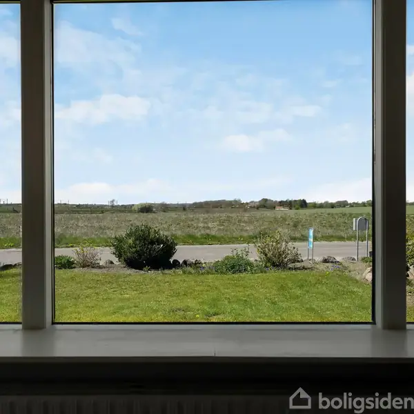 Et stort vindue viser en græsplæne og en vej i forgrunden med en åben mark og en blå himmel i baggrunden.