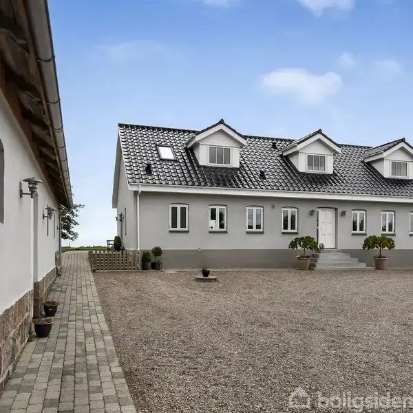 En hvid bygning med sort tag står i en gårdsplads med grus og fliser. En træplade med teksten "Bjerggaard" og nummer "26" ses på væggen til venstre.