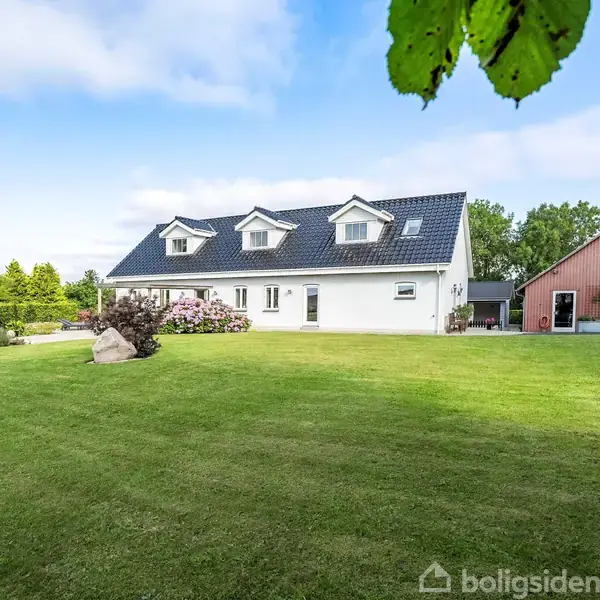 Et hvidt etplanshus med sort tag og tre kviste står på en grøn græsplæne med buske og træer i baggrunden. Til højre ses en rød udbygning.
