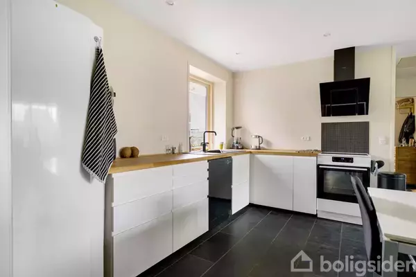 Hvidt køkken med sorte fliser og træbordplade. Rengøringsklud hænger til venstre. Moderne komfur og emhætte. Et vindue ved køkkenvasken giver naturligt lys.