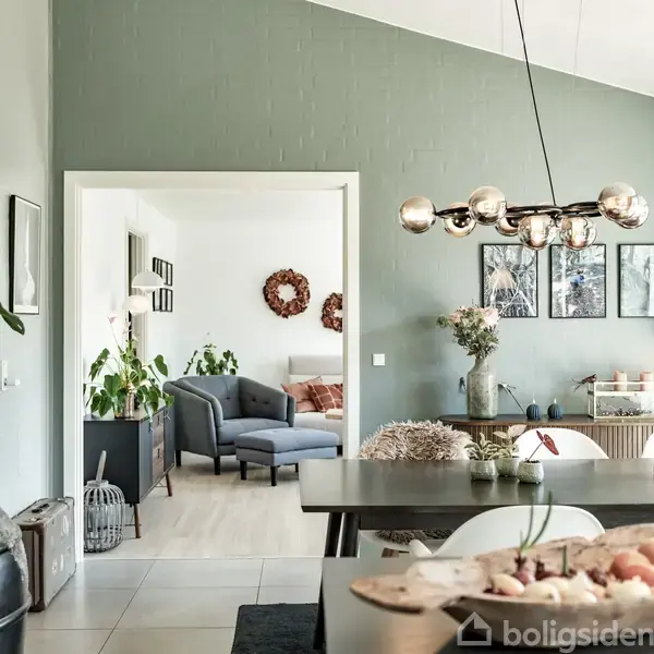 Spisebord i moderne stue med elegant lysekrone hængende over. Rummet er stilrent indrettet med grønne vægge og planter, og i baggrunden ses en tilstødende stue med sofa og dekorationer.