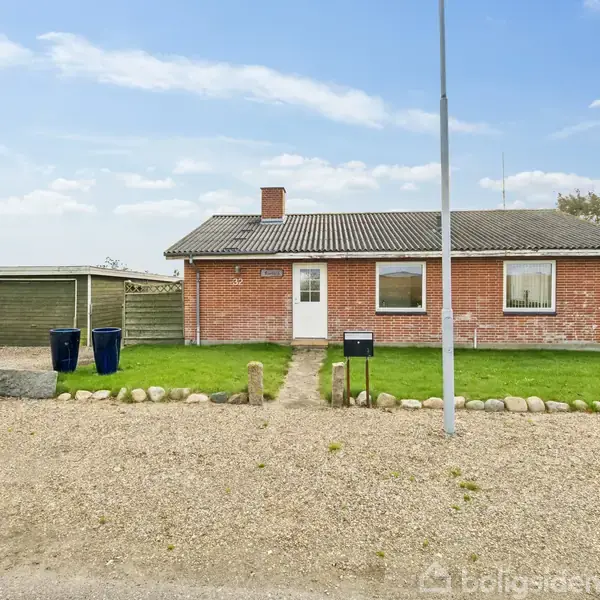 En enkelt-etagers rød murstensbygning står stille med en grusvej foran. En græsplæne og en række sten grænser til huset. Et halvtag og affaldsbeholdere ses til venstre.