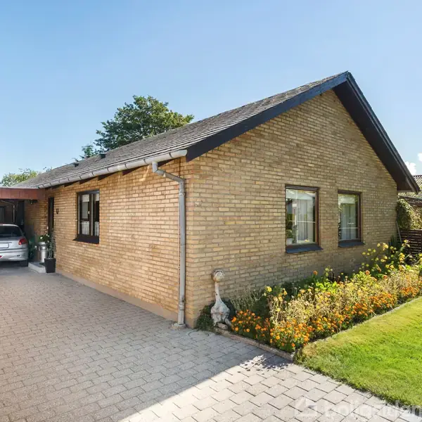 En murstensvilla i ét plan har en carport med en parkeret bil, omgivet af en velplejet have med blomster. Huset ligger i et solrigt boligområde.