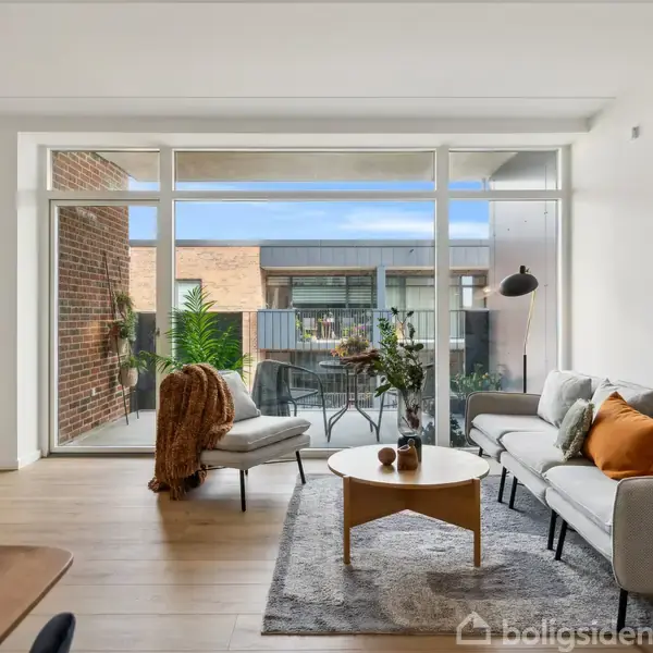 Stue med sofa, lænestol og sofabord på tæppe. Store vinduer åbner mod en altan med planter. Væggen har et kunstbillede markeret "KLEE." Lys og moderne indretning.