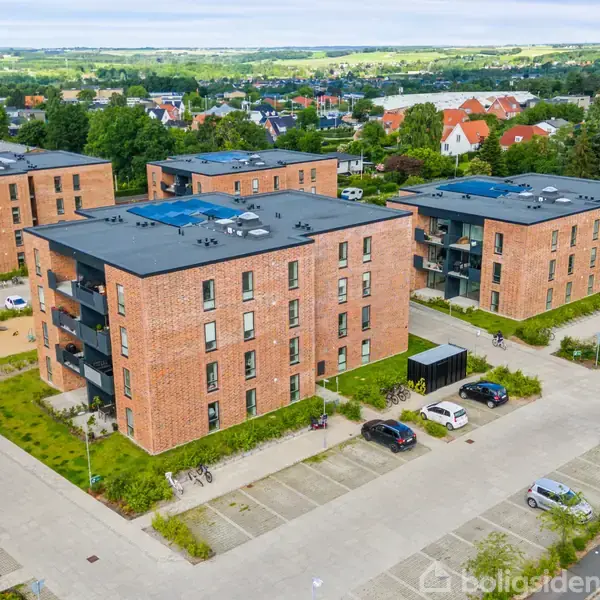 Flere rødstensbygninger står stille i et boligområde med parkerede biler omkring. Grønne områder og legeplads ses i baggrunden, mens landskabet breder sig mod horisonten.