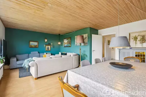 Stue med to grå sofaer og et tæppe på et trægulv, vægge i blå og hvid farve, udsmykket med malerier. Et spisebord med hvid dug står i forgrunden.
