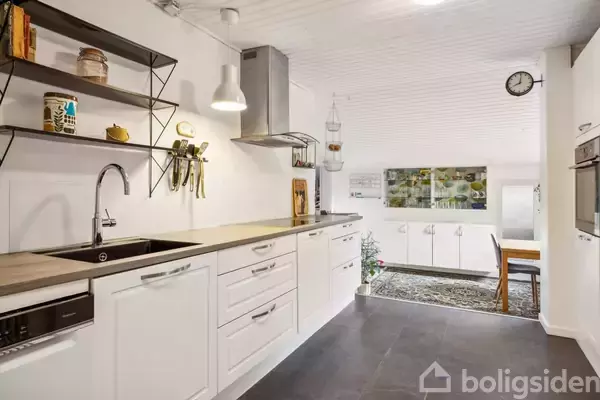 Køkken med hvide skabe og sort bordplade, moderne ovn og vask. En spiseplads med træbord ses i baggrunden, og en væg med hylder er til venstre. En rund væg-ur er til højre.