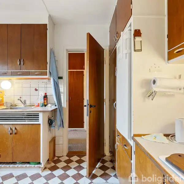 Køkken med brune træskabe og ternet gulv. Dør står åben mod et tilstødende rum. Køkkenudstyr og service er på bordpladen. Orange prikker dekorerer vægfliserne.