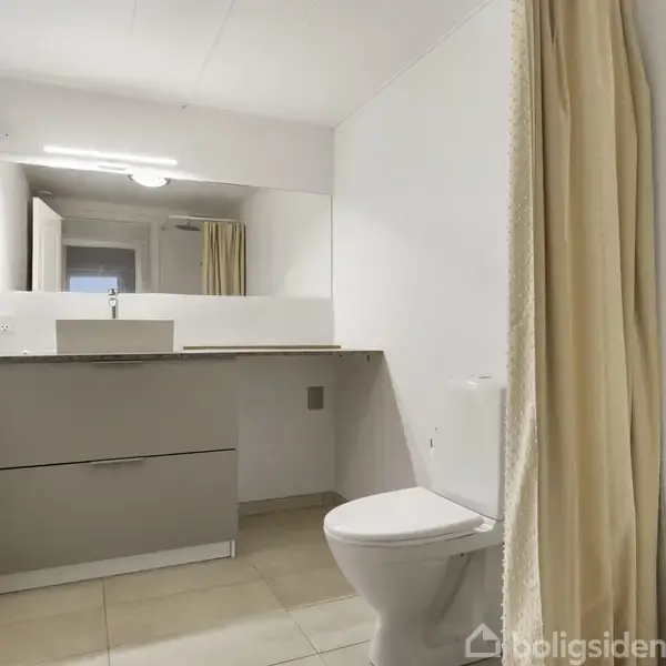 En moderne badeværelsesvask står på en grå skabsenhed. En hvid toiletkumme er placeret til højre. Et brusehoved og et beige forhæng er på højre side.