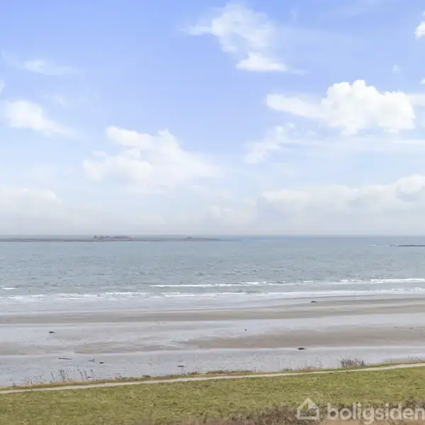 Strand strækker sig langs kysten med bølger der rammer kystlinjen. I baggrunden ses havet og en horisont under en let overskyet himmel. Ingen tekst til stede.