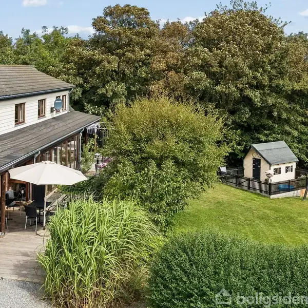 Hus med to etager står ved en terrasse med parasol. I haven ses et legehus og gyngestativ omgivet af græsplæne og buske, med træer i baggrunden.