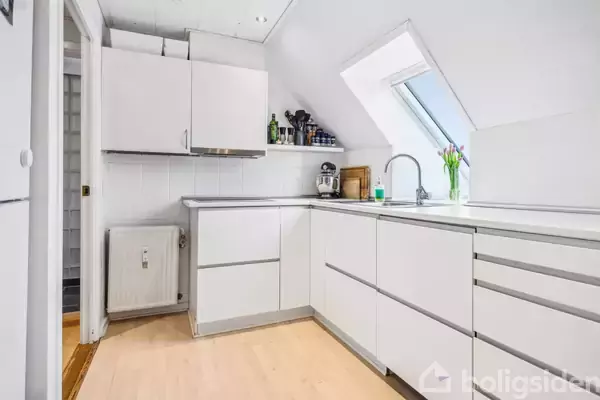 Et moderne køkken med hvide skabe og en skrå kvistvindue. En vase med tulipaner står ved en håndvask. Flasker og en kaffemaskine er på bordpladen.