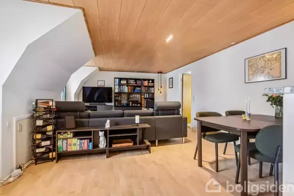 Sofaer står i stue med fladskærms-TV foran reol fyldt med bøger. Ved siden af er et spisebord med fire stole. Loftet er i træ.