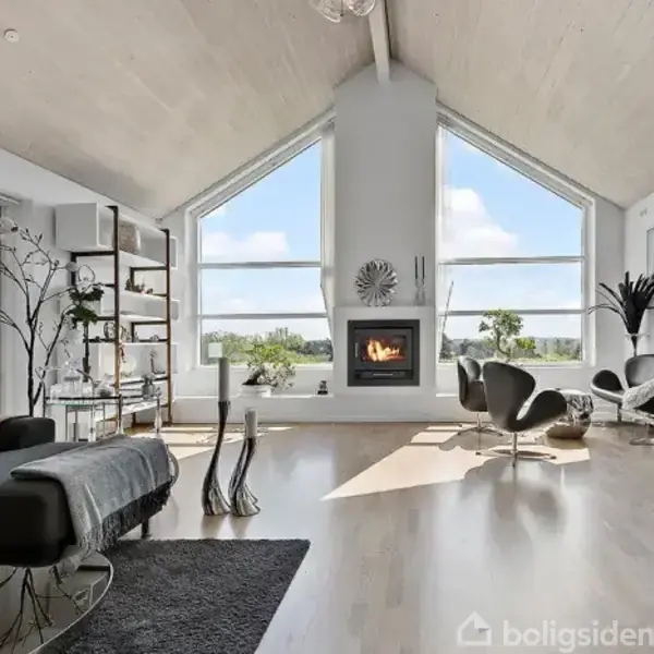 Stue med moderne møbler, hvor en pejs brænder. Store vinduer slipper lys ind. Rummet har minimalistisk dekoration med grønne planter og en stilfuld sort sofa.