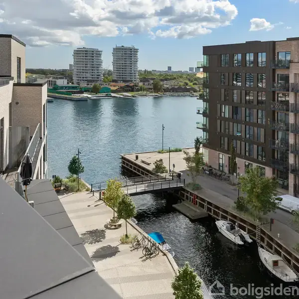 Bygninger omgiver en kanal med en bro i forgrunden. En lystbåd er ved kajen, og moderne højhuse ses i baggrunden. Vandet reflekterer omgivelserne i dagslys.