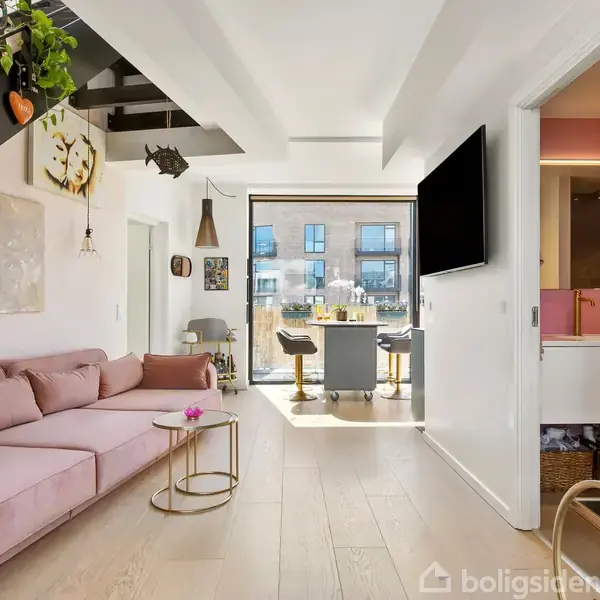 Rosa sofa står i en stue med lyst trægulv, hængende planter og moderne kunst. Et stort vindue fører til en balkon, mens et tv hænger på væggen ved siden af et åbent køkken.