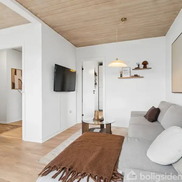 Grå sofa med brune puder står ved hvide vægge. Rundt sofabord med lamper og dekoration. Væghængt TV i elegant, minimalistisk stue med trægulv og loft.