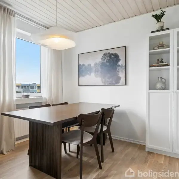 Et spisebord med fire stole står i et lyst rum ved siden af et højt skab. En lampe hænger over bordet. På væggen hænger et landskabsmaleri, og vinduet har hvide gardiner.