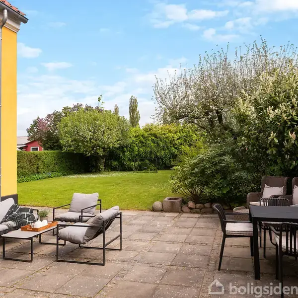 En terrasse med stole og bord står på fliser ved siden af et gulmalet hus. Den omkringliggende have er grøn og frodig med buske og træer, der skaber en afslappende atmosfære.