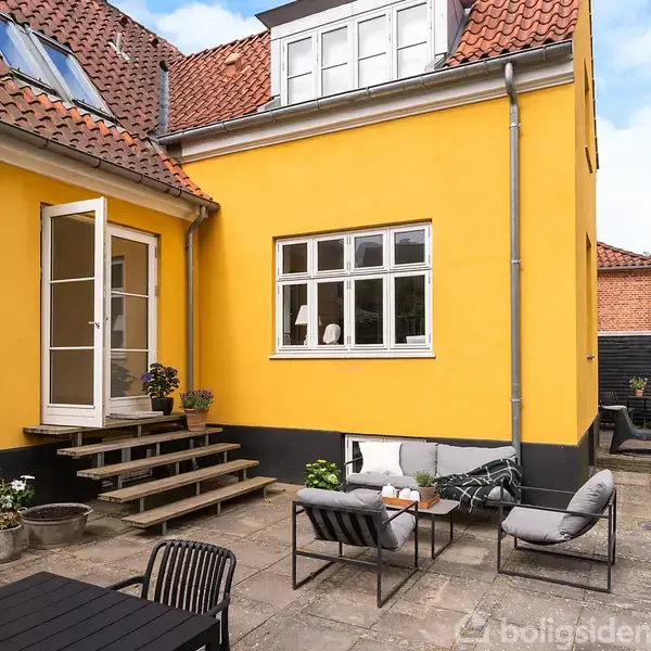 Gul murstensbygning med terrasse, hvor trapper fører til en glasdør. På terrassen står havemøbler og potteplanter. Den omkringliggende atmosfære er hyggelig og privat.