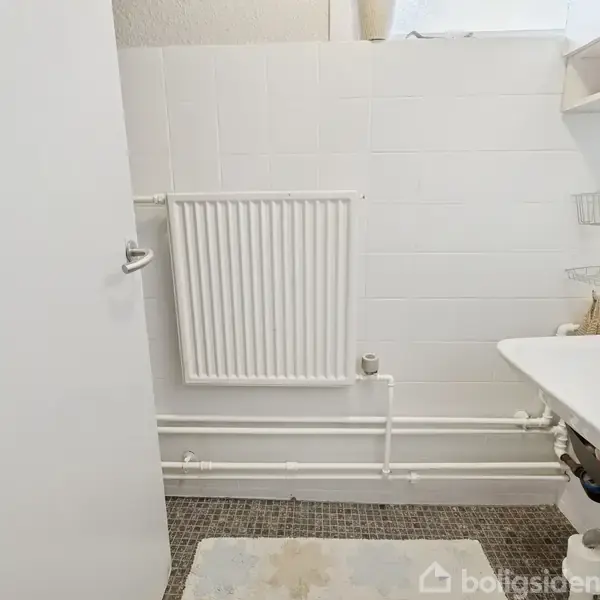 En hvid radiator hænger på en badeværelsesvæg. Ved siden af er der en hvid vask med tilbehør som sæbedispenser og tandkrus. Flisegulvet er dækket af en lille måtte.