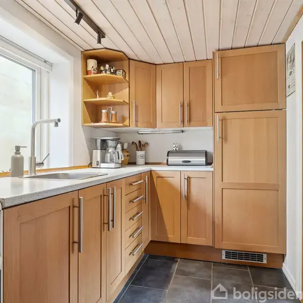 Køkken med træskabe, vask og apparater på bordplade, belysning i loftet. Rummet har et vindue mod venstre og flisegulv, omgivet af hvide vægge og en trædør ind til et andet rum.