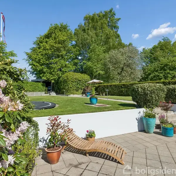 Træliggestol står på flisebelagt terrasse, omgivet af blomsterkrukker. I baggrunden ses en grøn have med hække, træer og en trampolin. Solskin understreger den fredfyldte atmosfære.
