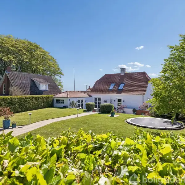 En hvid villa med rødt tegltag står i en velplejet have med plæne og flagstang. Omkring huset er der træer og en trampolin. Solrigt vejr under en klar blå himmel.