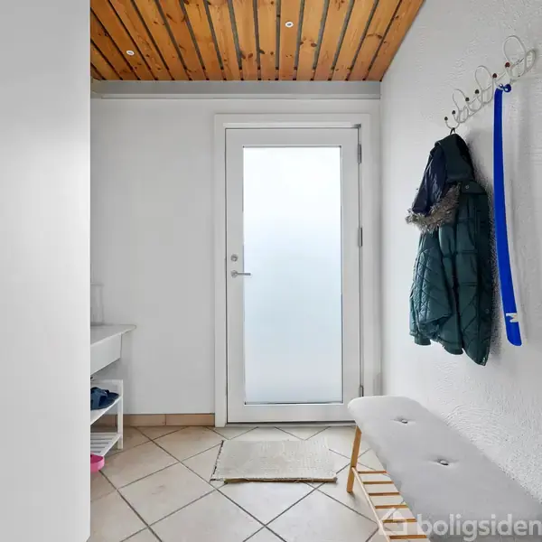 En entre med klinkegulv og hvidmalede vægge. Dør i baggrunden, bænk til højre, jakker og blå snor på knagerække. Hvidt skab til venstre. Træloft.