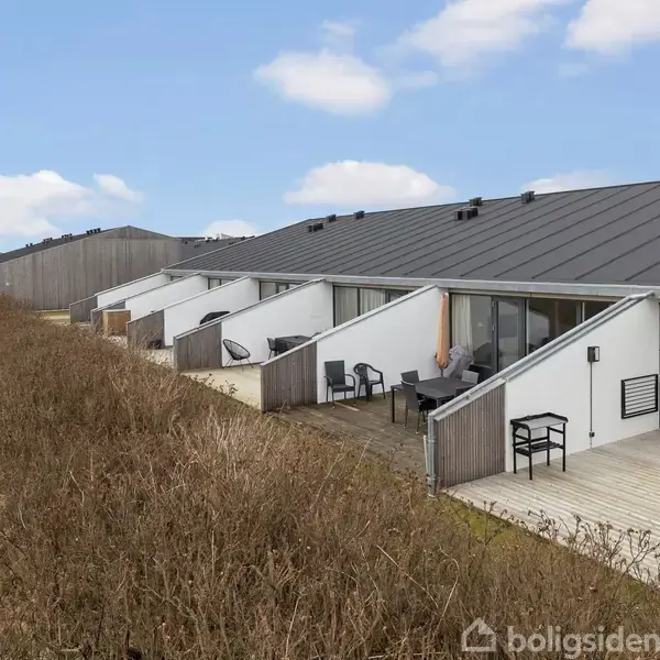 Rækkehuse med skrå tage står på række. Terrasser med havemøbler vender ud mod en åben græsplæne. Tæt hæk afgrænser ejendommens kant. Skyfri blå himmel i baggrunden.